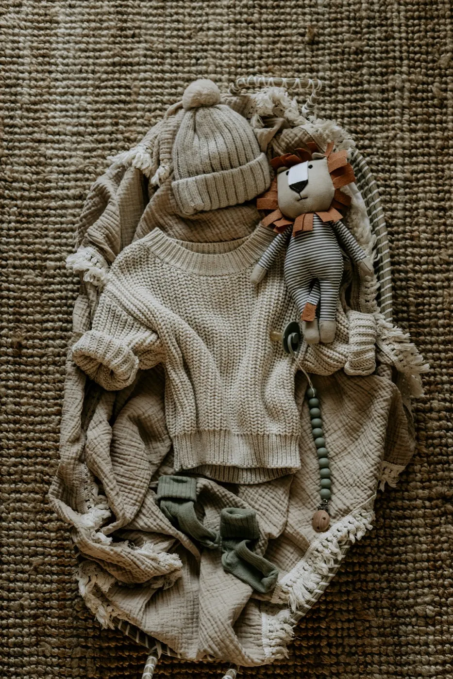A flatlay on a jute rug of a cream chunky knit baby sweater, a matching ribbed pompom beanie, a crinkle muslin blanket, sage socks, a striped lion soft toy and a pacifier clip