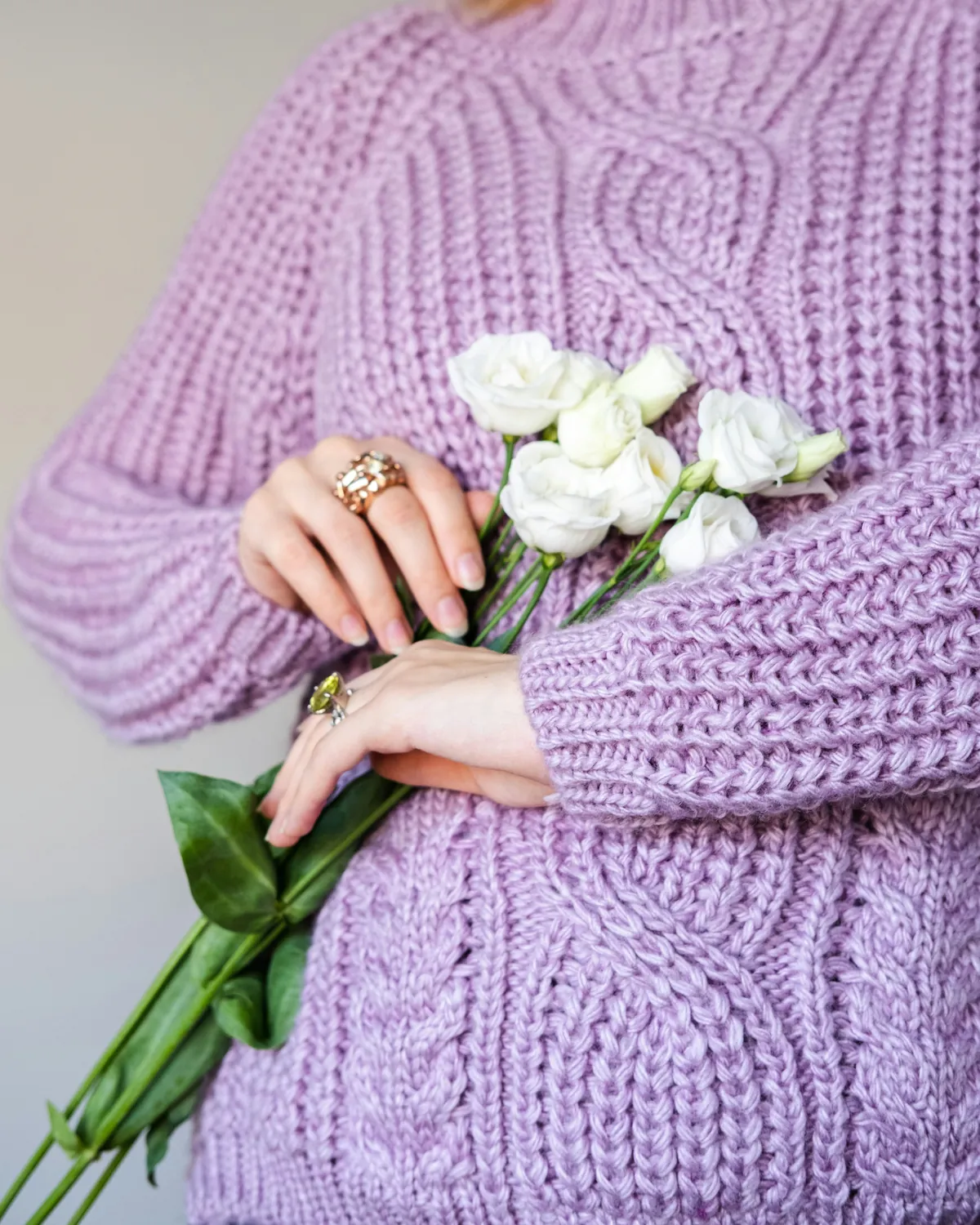 A woman, head out of frame, in a lilac chunky rib-and-cable knit sweater, holding a small bunch of white lisianthus