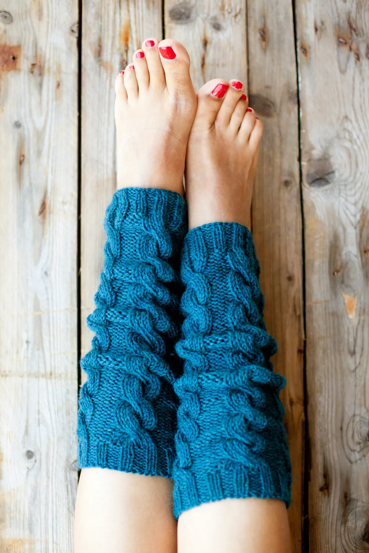 A pair of teal cable-knit leg warmers on bare lower legs, against weathered grey planks