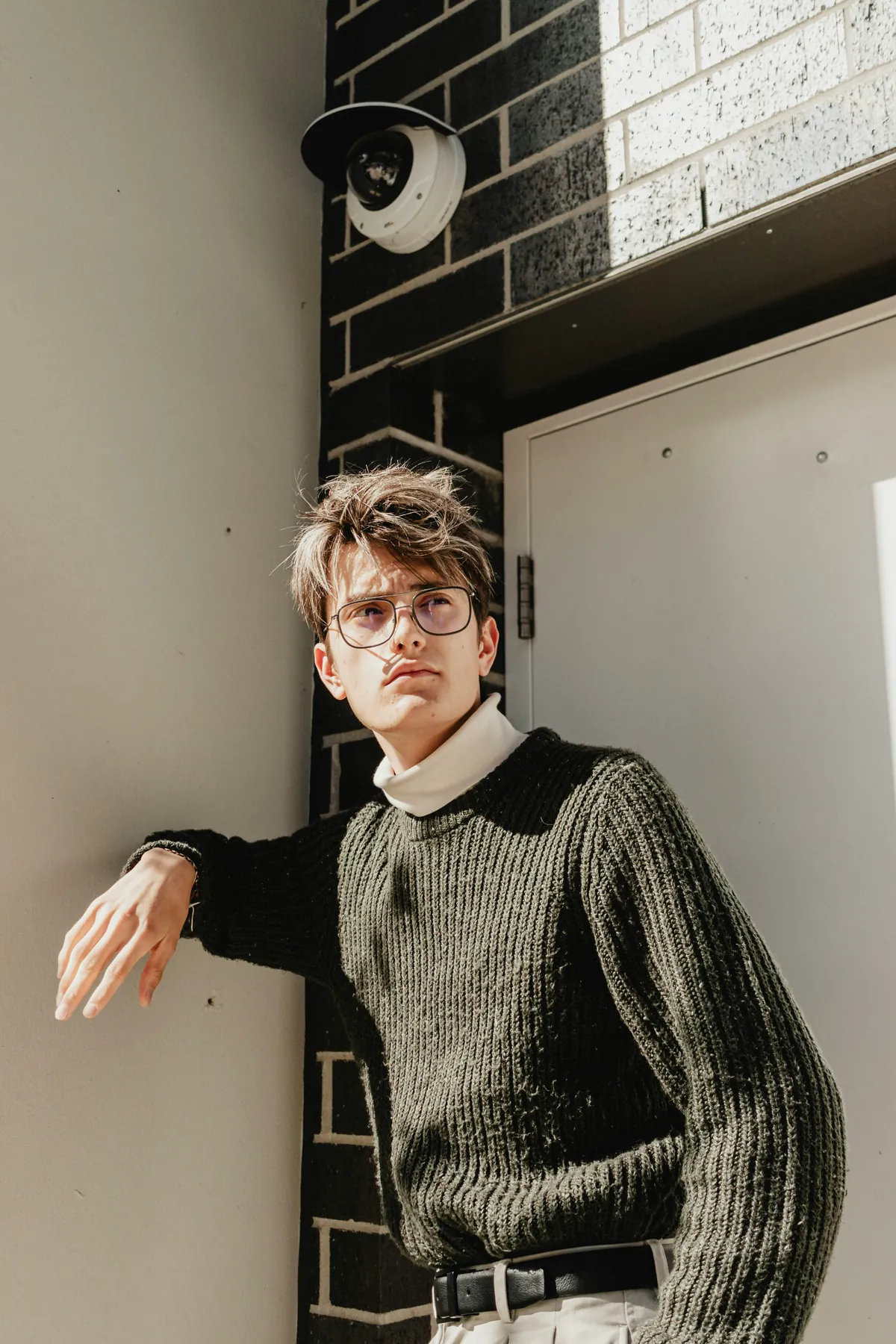 A young man in a dark olive raglan knit sweater over a white turtleneck, leaning against a brick wall outdoors