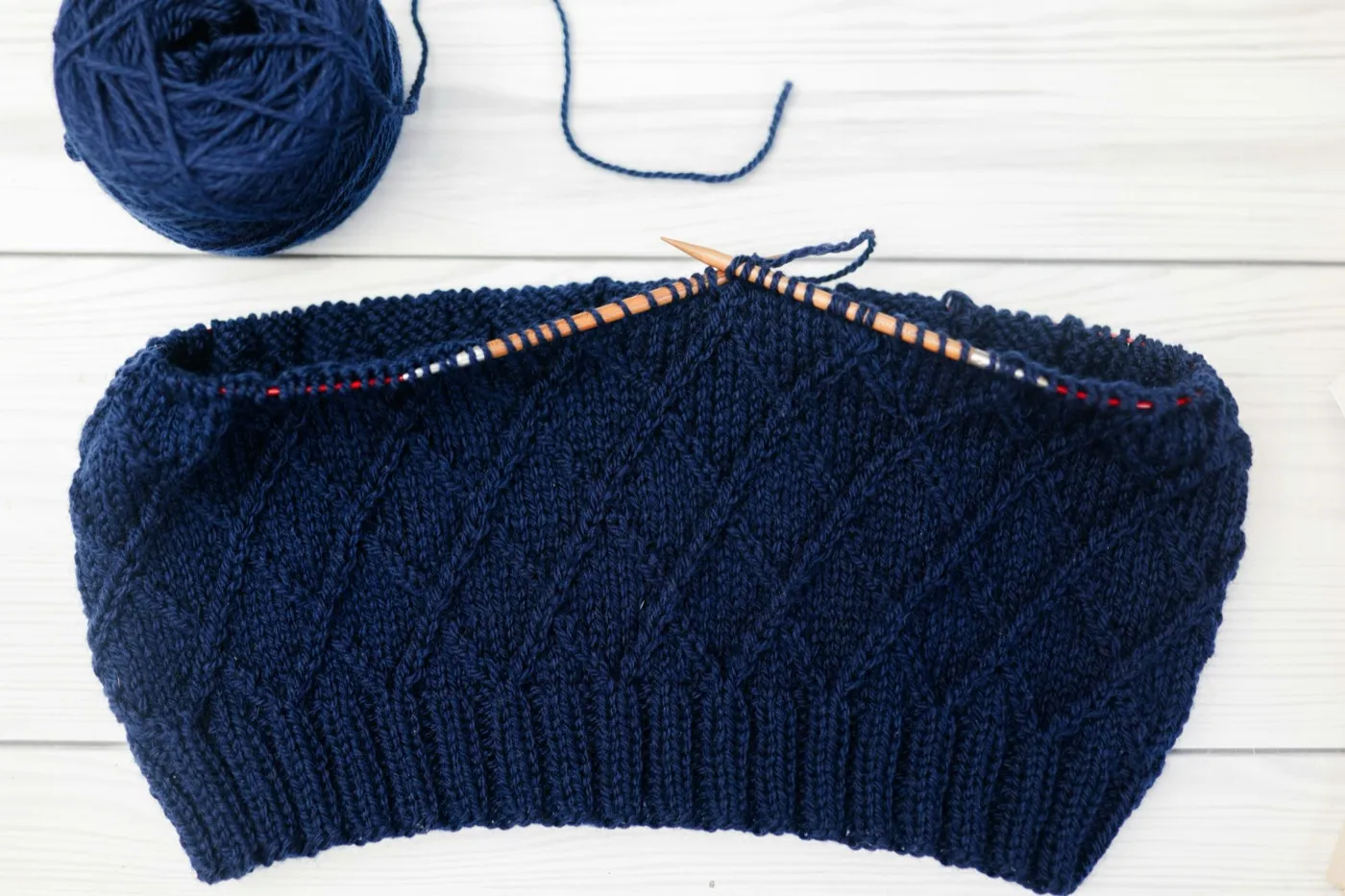 A navy in-progress knitted panel on wooden circular needles, beside a ball of navy yarn on white painted planks