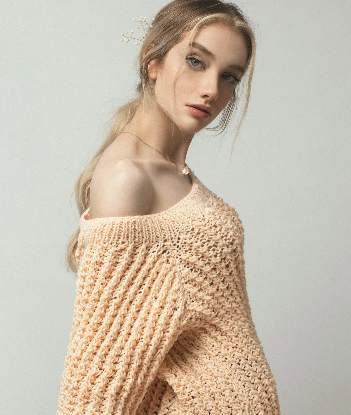 A woman in a blush chunky off-shoulder knit sweater, gypsophila in her hair, against a grey studio backdrop