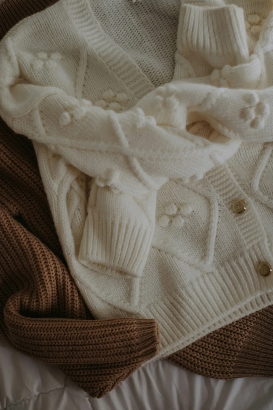 An ivory knit cardigan with raised bobble-flower motifs and tortoiseshell buttons, laid flat on a bed beside a rust-brown ribbed knit