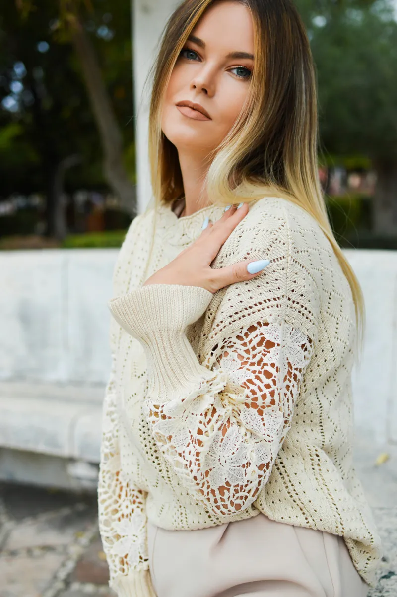 A woman in a cream open-work sweater with crocheted lace panels, outdoors beside a pale stone wall