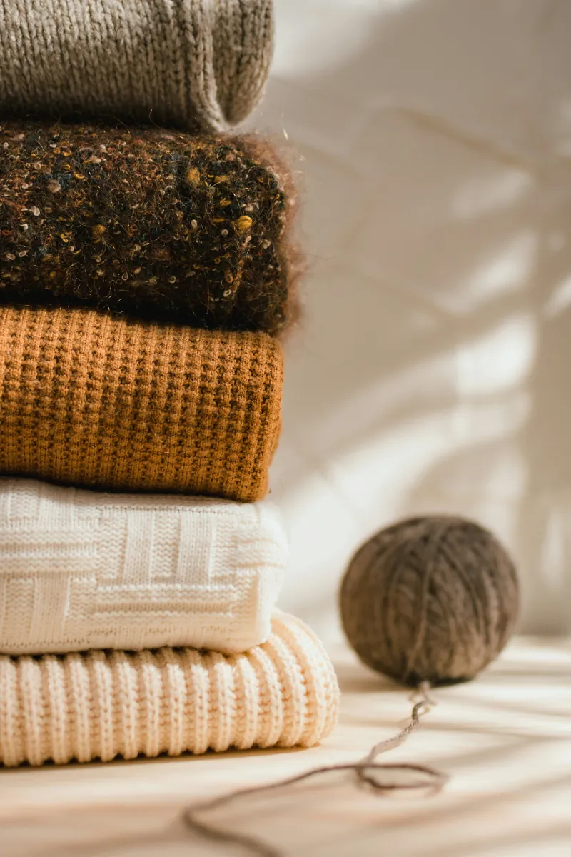 A stack of five folded knit sweaters in grey, brown tweed, mustard, ivory and cream, beside a single ball of wound grey-brown yarn with a loose thread trailing across a sunlit wooden surface